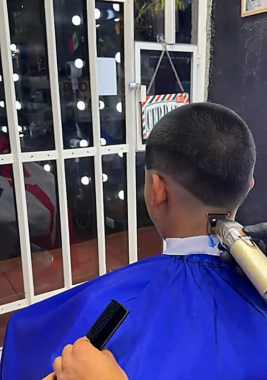Mano tatuada del barbero perfilando una nuca con la máquina, frente al ventanal con rejas blancas